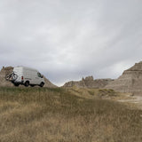 White Sprinter van with bike rack parked in front of striking badlands formations. Ideal for an RV rental road trip!