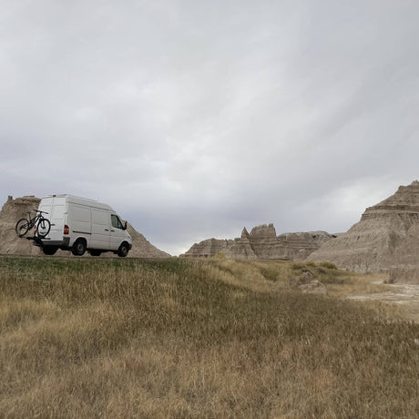 White Sprinter van with bike rack parked in front of striking badlands formations. Ideal for an RV rental road trip!