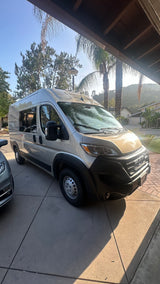 A gold Ram ProMaster camper van, ready for an RV rental, parked on a concrete driveway under a clear sky.