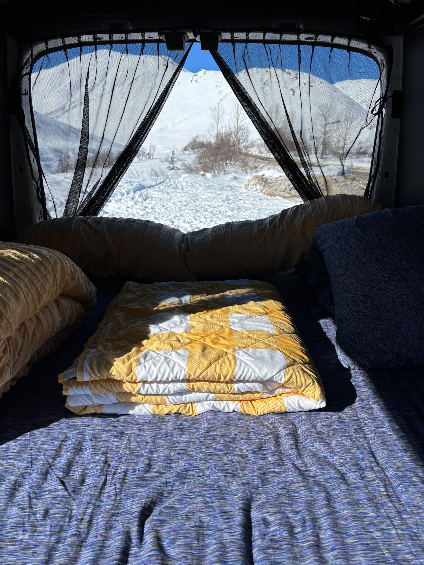 Cozy RV rental bed with yellow and blue blankets facing snowy mountains framed by black mesh curtains.