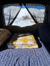 Cozy RV rental bed with yellow and blue blankets facing snowy mountains framed by black mesh curtains.