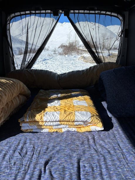Cozy RV rental bed with yellow and blue blankets facing snowy mountains framed by black mesh curtains.