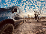 Dark adventure van reflects a vibrant cloudy sky, parked in a desert with sparse trees. Perfect RV rental escape!