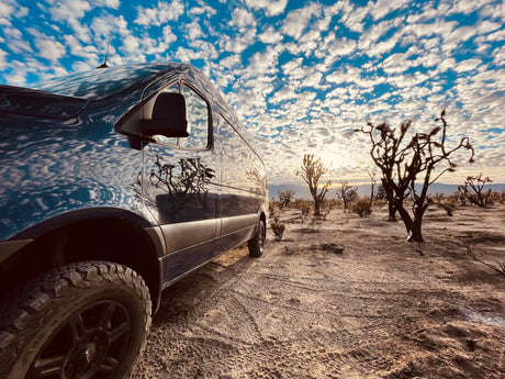 Dark adventure van reflects a vibrant cloudy sky, parked in a desert with sparse trees. Perfect RV rental escape!