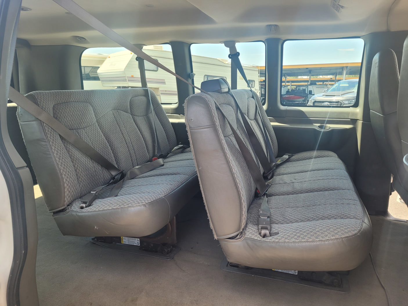 View inside an RV rental shuttle van, showing two rows of grey fabric seats with seatbelts, and RVs visible outside.