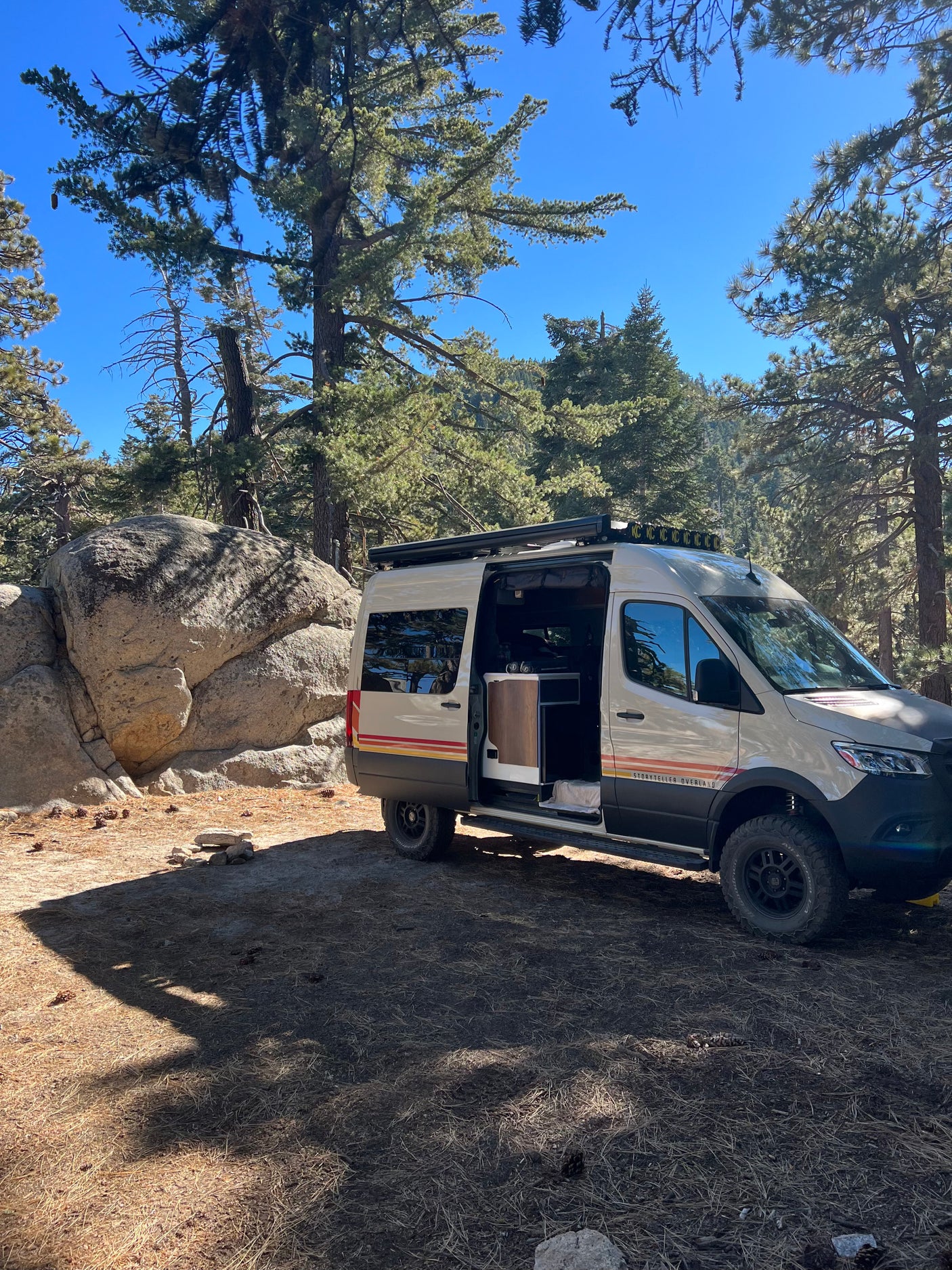 A robust Storyteller Overland camper van, ideal for rv rental, sits in a sunlit forest campsite among large rocks.