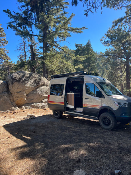 A robust Storyteller Overland camper van, ideal for rv rental, sits in a sunlit forest campsite among large rocks.