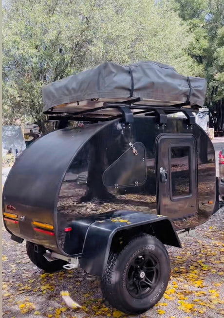 Black teardrop RV rental camper with rooftop tent, on large tires, parked amidst yellow leaves under trees for your next adventure.