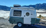 An RV rental: a white & green camper with Dometic AC & Fiamma awning, parked near snowy mountains under a clear sky.