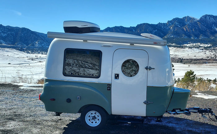 An RV rental: a white & green camper with Dometic AC & Fiamma awning, parked near snowy mountains under a clear sky.