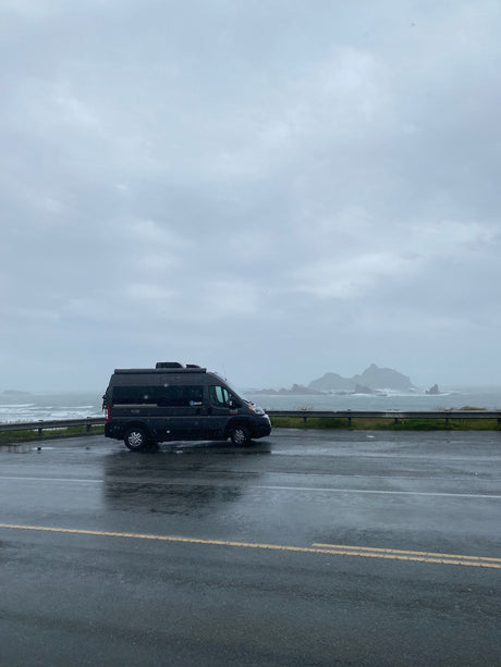 Dark RV rental parked on a wet coastal road, overlooking a stormy ocean with distant rocky islets under a grey sky.