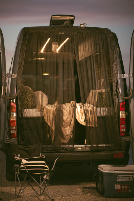 Cozy RV rental setup at twilight. Back of camper van open, revealing a lit bed protected by a net. Camp chair & cooler outside.