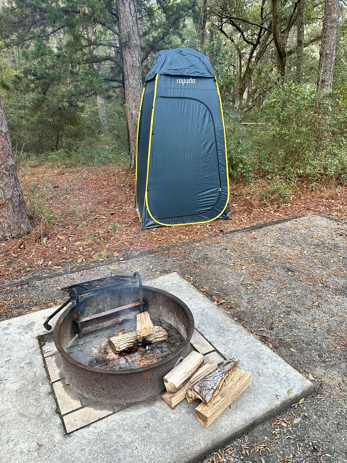 An RV rental campsite with a metal fire pit containing logs and smoke, alongside a tall grey privacy tent in a forest.