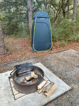 An RV rental campsite with a metal fire pit containing logs and smoke, alongside a tall grey privacy tent in a forest.