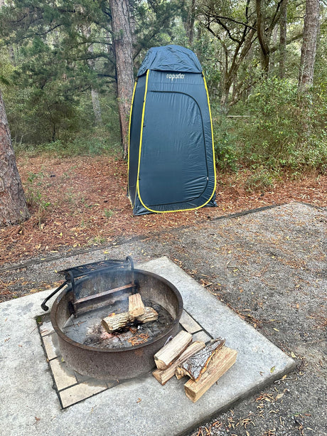 An RV rental campsite with a metal fire pit containing logs and smoke, alongside a tall grey privacy tent in a forest.