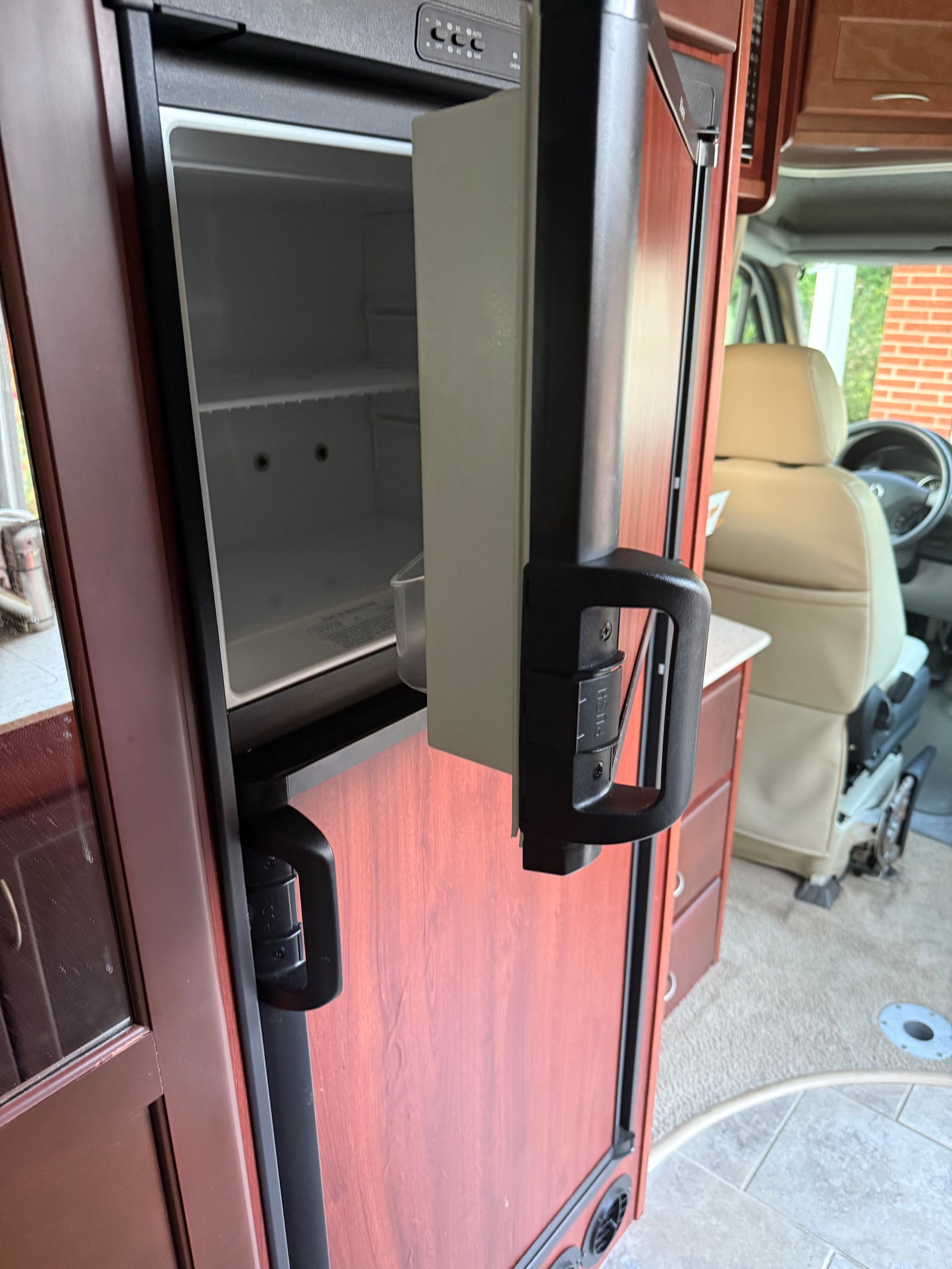 Inside an RV rental: empty refrigerator, top controls (DC/AC/GAS), and front cabin beyond. Compact travel features.