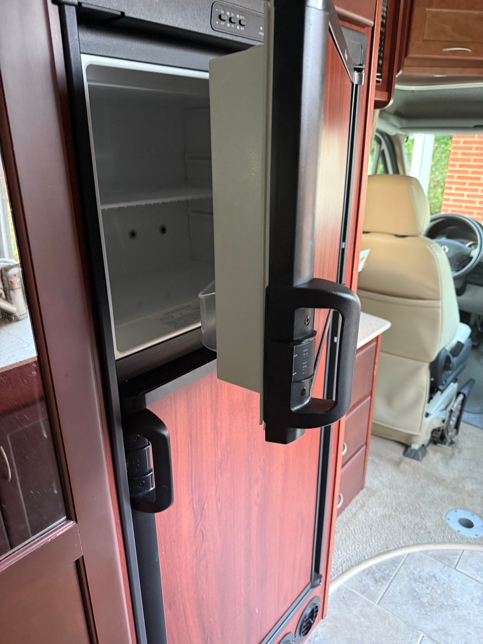 Inside an RV rental: empty refrigerator, top controls (DC/AC/GAS), and front cabin beyond. Compact travel features.