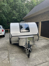 Compact white travel trailer, ideal for an rv rental adventure, parked in a driveway. Features a large front window & storage box.