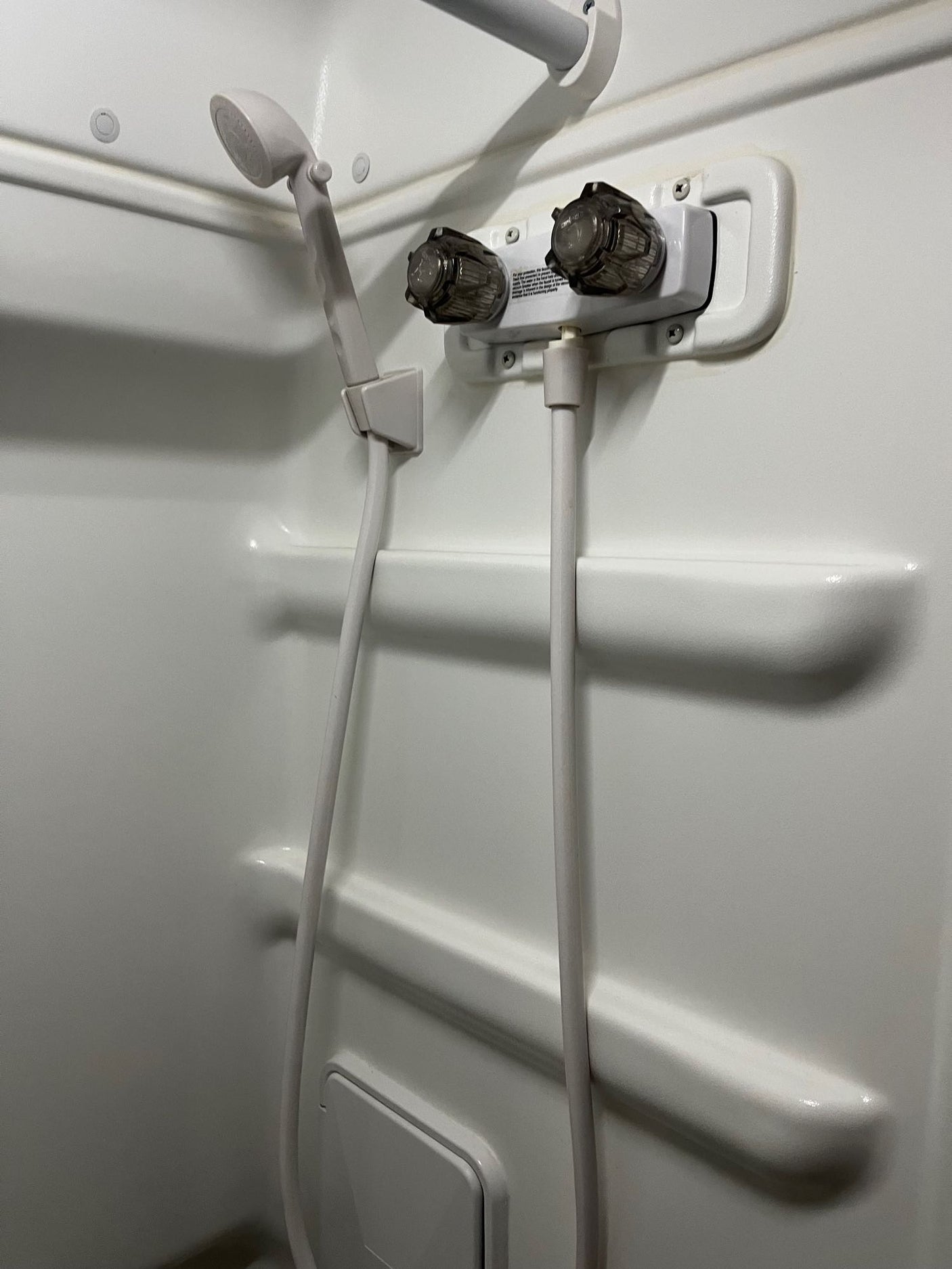 Interior of a compact white RV rental shower with handheld shower head, hot/cold faucet, and integrated shelving.