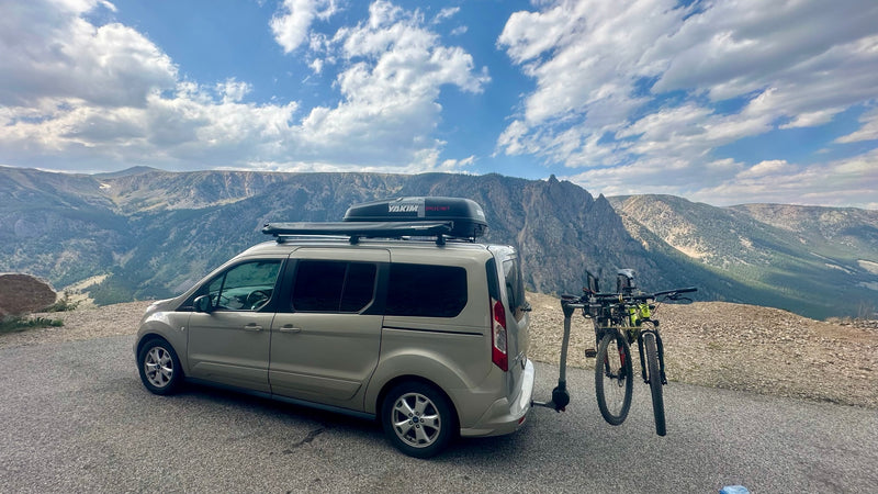 This RV rental van, loaded with bikes, roof cargo box, and awning, sits on a mountain road under a cloudy sky, ready for adventure. — photo 14