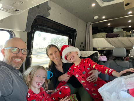 A smiling family of four in a cozy RV rental van, decorated with snowflakes. Kids wear Christmas pajamas and a Santa hat.