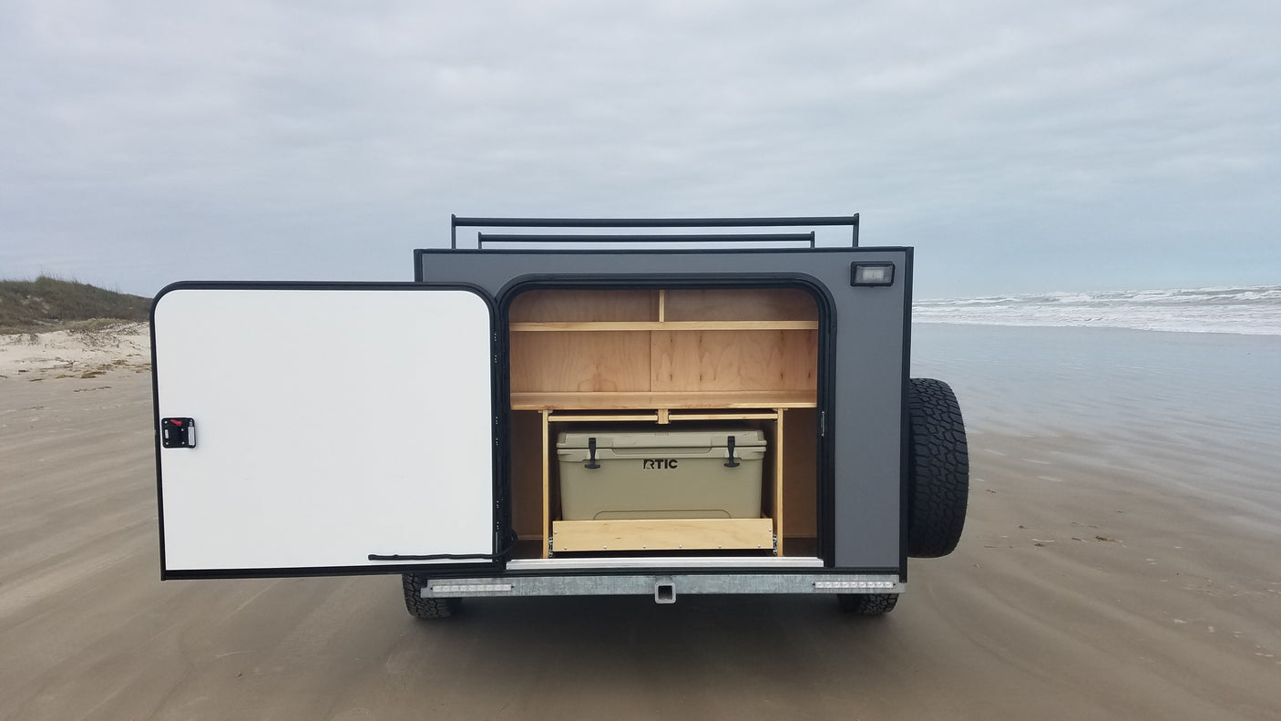 Open off-road camper trailer (RV rental) on a beach. Rear shows organized storage shelves & RTIC cooler on pull-out for trips.