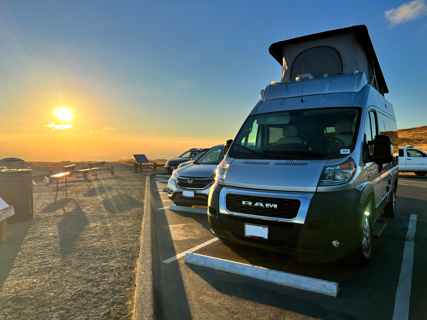 Sunset view from a scenic overlook featuring a parked RV rental with a pop-top roof and other vehicles.