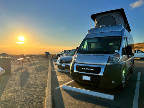 Sunset view from a scenic overlook featuring a parked RV rental with a pop-top roof and other vehicles.