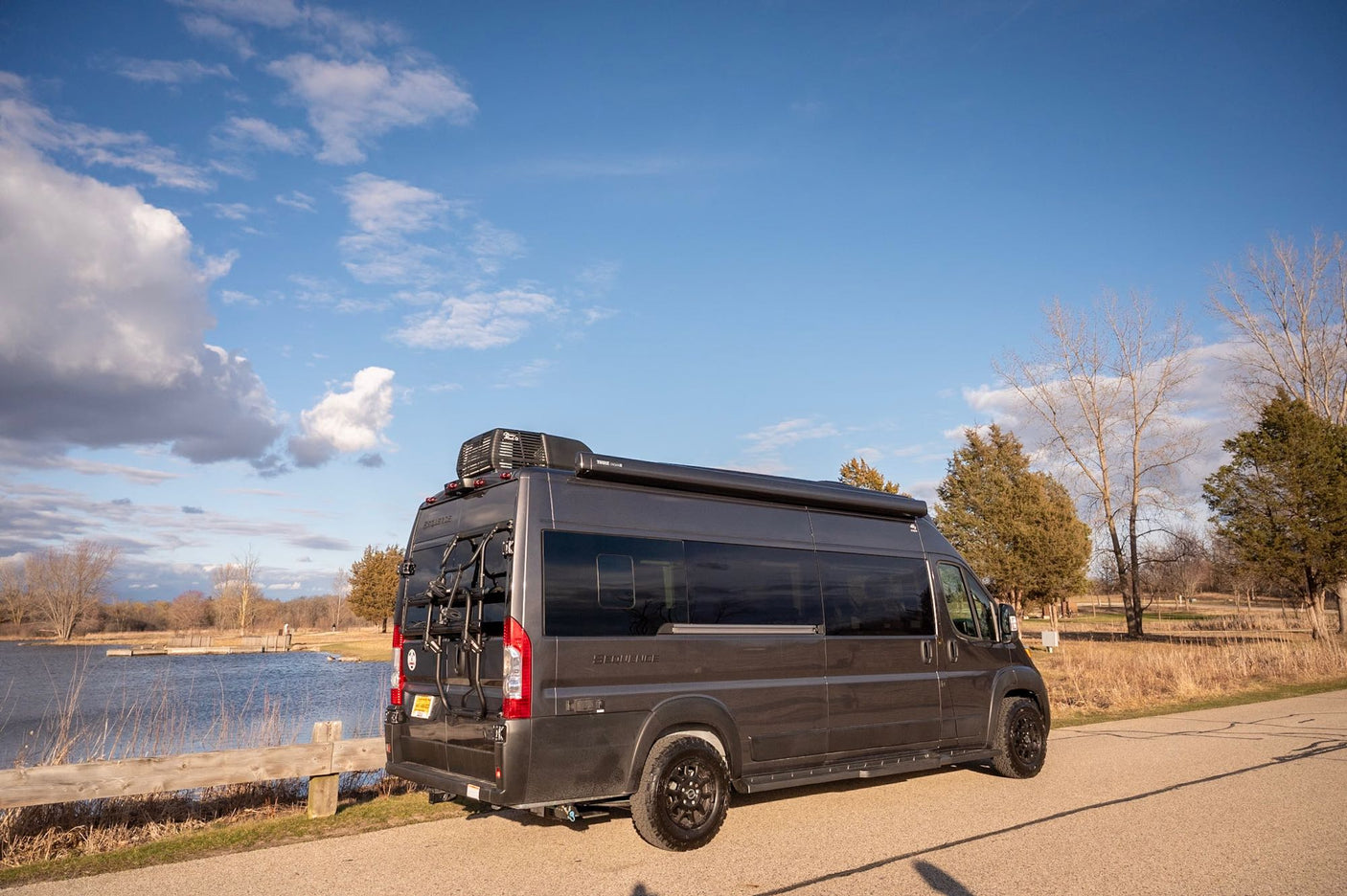 Dark grey camper van RV rental by a lake. Equipped with a bike rack & awning for outdoor adventures.