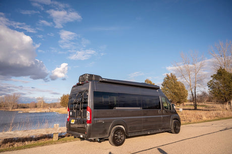 Dark grey camper van RV rental by a lake. Equipped with a bike rack & awning for outdoor adventures.