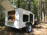 White mini teardrop camper RV rental with an open rear kitchen full of snacks, parked in a sunny forest setting.
