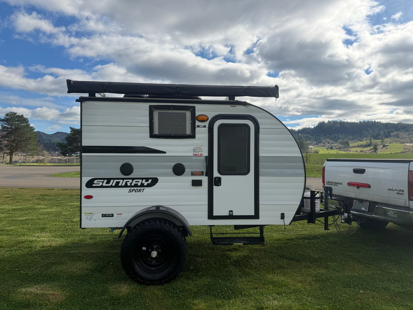 Sunray Sport RV rental: a white and gray teardrop camper trailer hitched to a truck, ready for adventure by a scenic lake and hills.