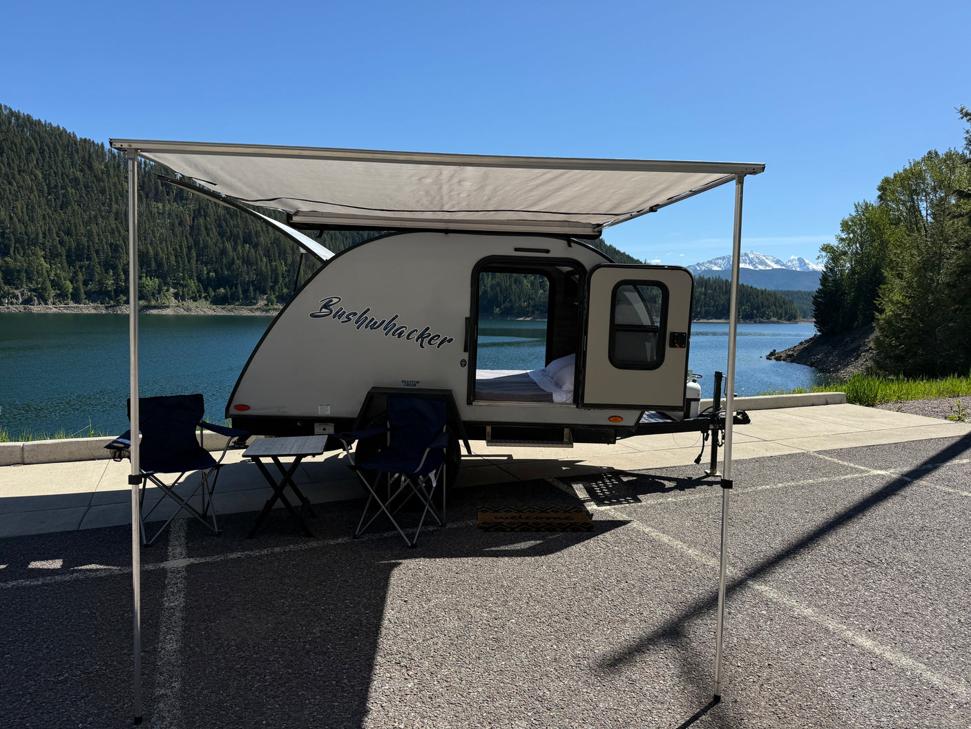 Serene rv rental setup: a "Bushwhacker" teardrop camper with an awning, chairs, by a lake and snowy mountains.