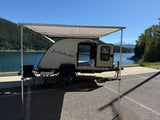 Serene rv rental setup: a "Bushwhacker" teardrop camper with an awning, chairs, by a lake and snowy mountains.