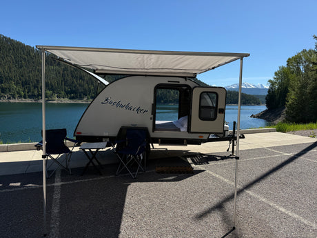 Serene rv rental setup: a "Bushwhacker" teardrop camper with an awning, chairs, by a lake and snowy mountains.