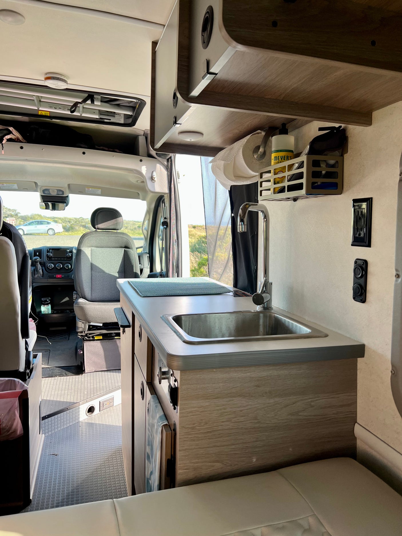RV rental interior: compact kitchen with sink, paper towels, cleaning supplies, driver's cab, and seating.