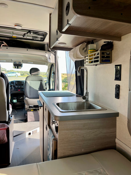 RV rental interior: compact kitchen with sink, paper towels, cleaning supplies, driver's cab, and seating.