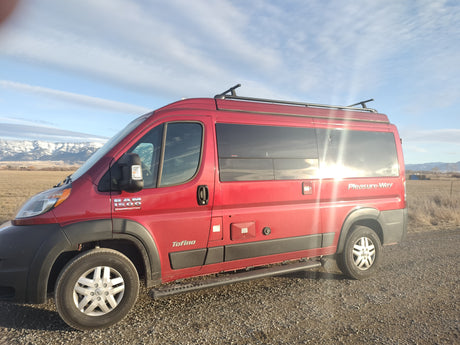 Red Pleasure-Way Tofino camper van, an ideal RV rental, parked on a gravel road with snowy mountains in the background.