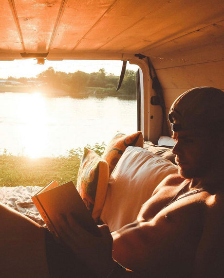 Man relaxing in an RV rental, reading a book by the river at sunset. Cozy van life scene.