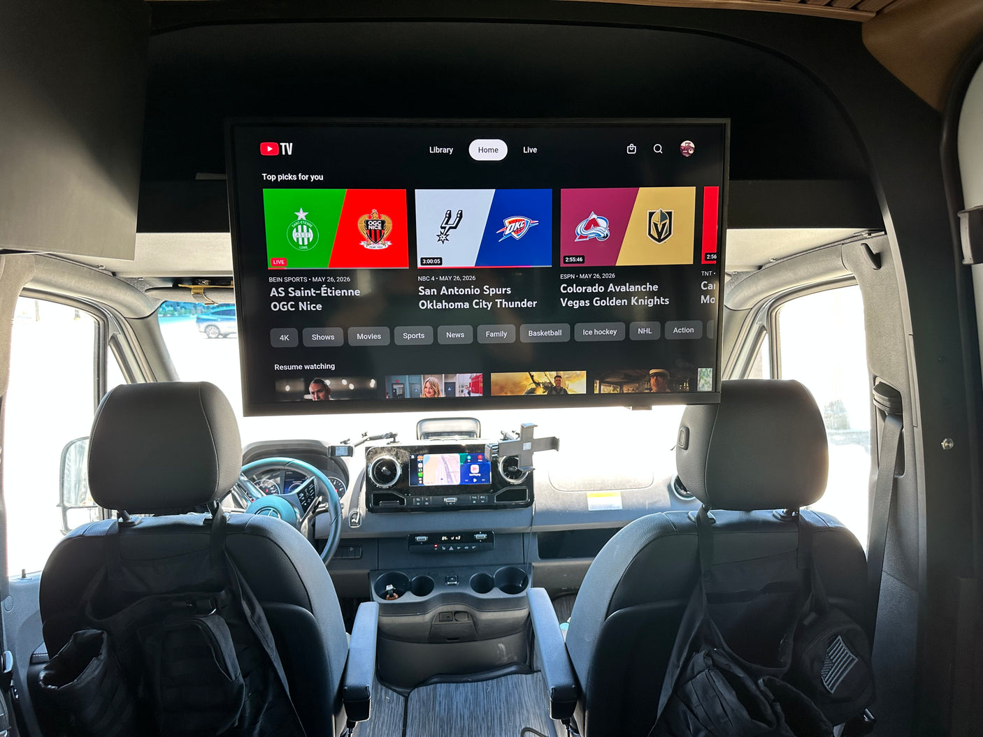 RV rental interior: A large TV above the dash displays YouTube TV with live sports. Front seats and vehicle controls visible.