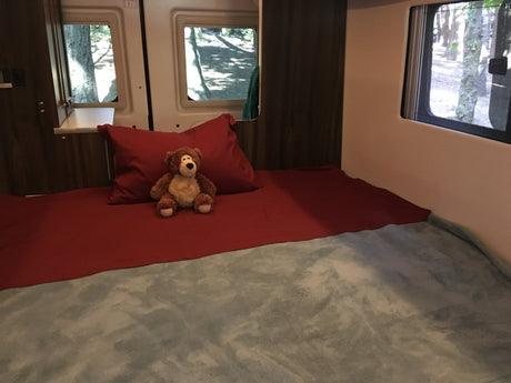 An RV rental's cozy bed features red linens, a teddy bear, and window views of nature.