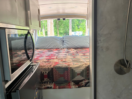 Explore this rv rental interior: a cozy bed with quilt, microwave, and a shower corner, set for relaxing road trips.