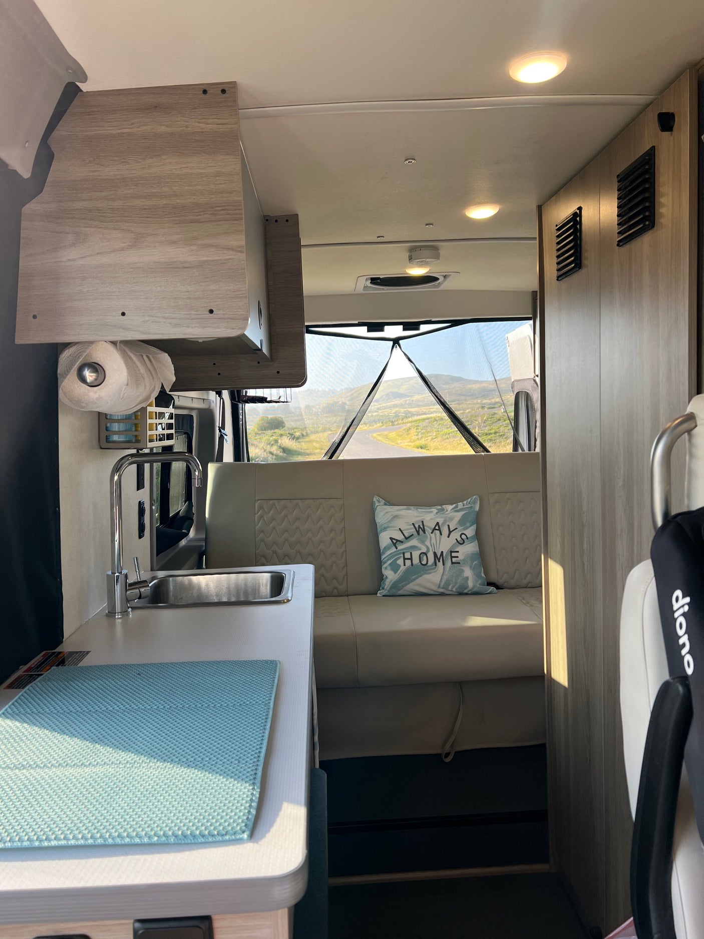 Cozy RV rental interior with kitchen sink, seating area, and a pillow reading "ALWAYS HOME." Scenic view through the rear screen.
