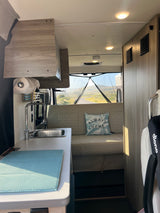 Cozy RV rental interior with kitchen sink, seating area, and a pillow reading "ALWAYS HOME." Scenic view through the rear screen.