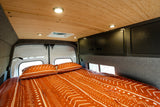 Comfortable RV rental bed with patterned bedding, wood ceiling, overhead cabinets, and functional lighting.