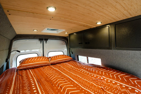 Comfortable RV rental bed with patterned bedding, wood ceiling, overhead cabinets, and functional lighting.