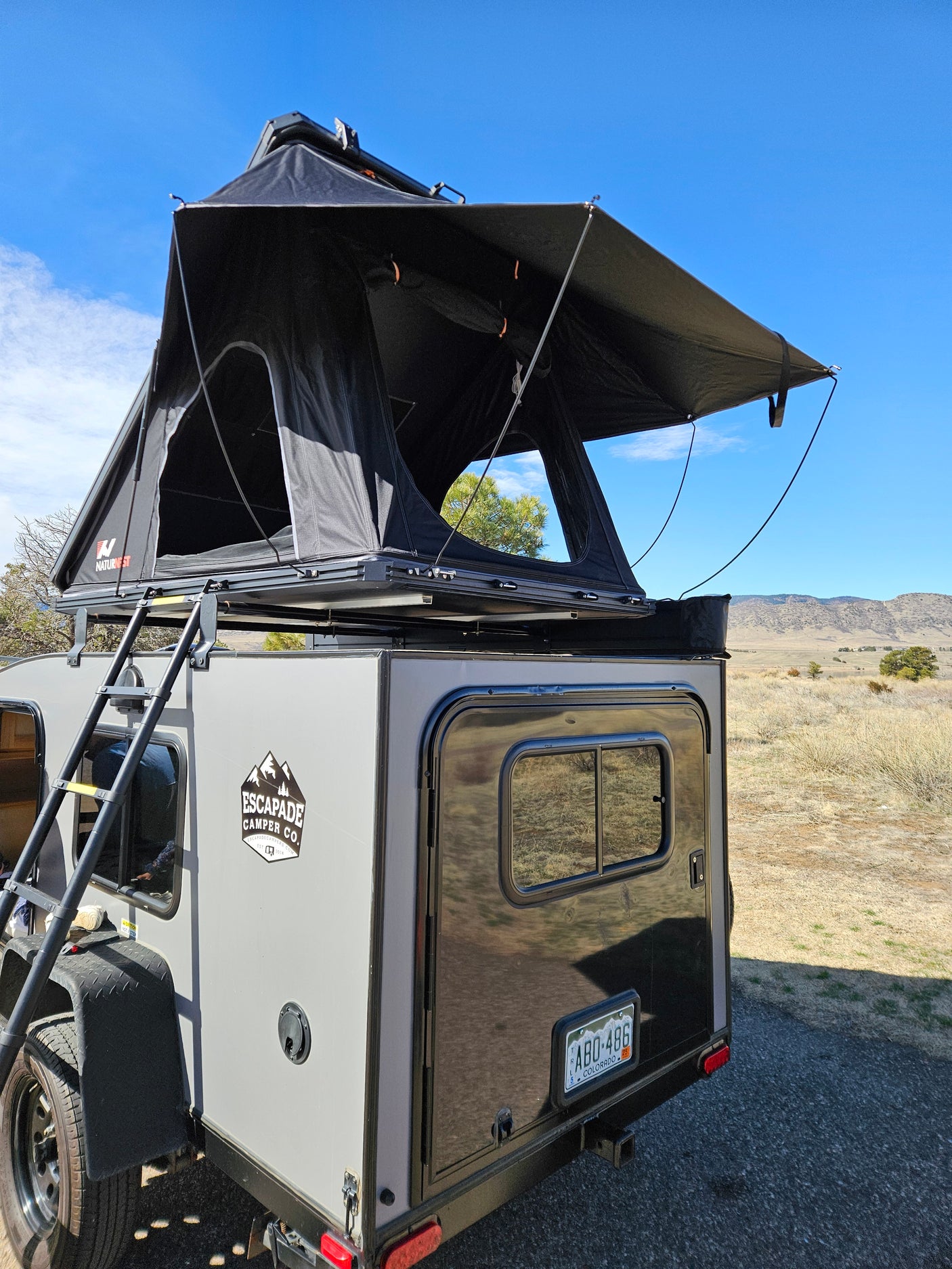Compact RV rental: A grey Escapade Camper with a black NatureNest rooftop tent deployed in a scenic Colorado landscape.