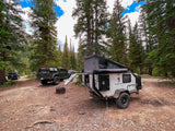 A pop-top Mission Overland trailer and Jeep Gladiator are set up for an outdoor RV rental adventure in a forest.