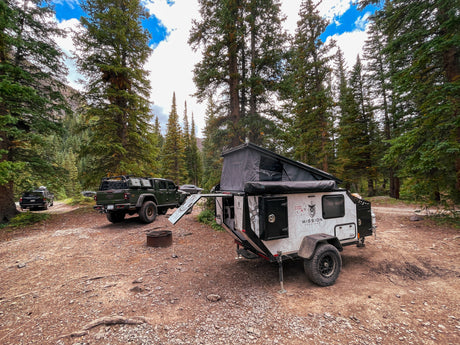 A pop-top Mission Overland trailer and Jeep Gladiator are set up for an outdoor RV rental adventure in a forest.
