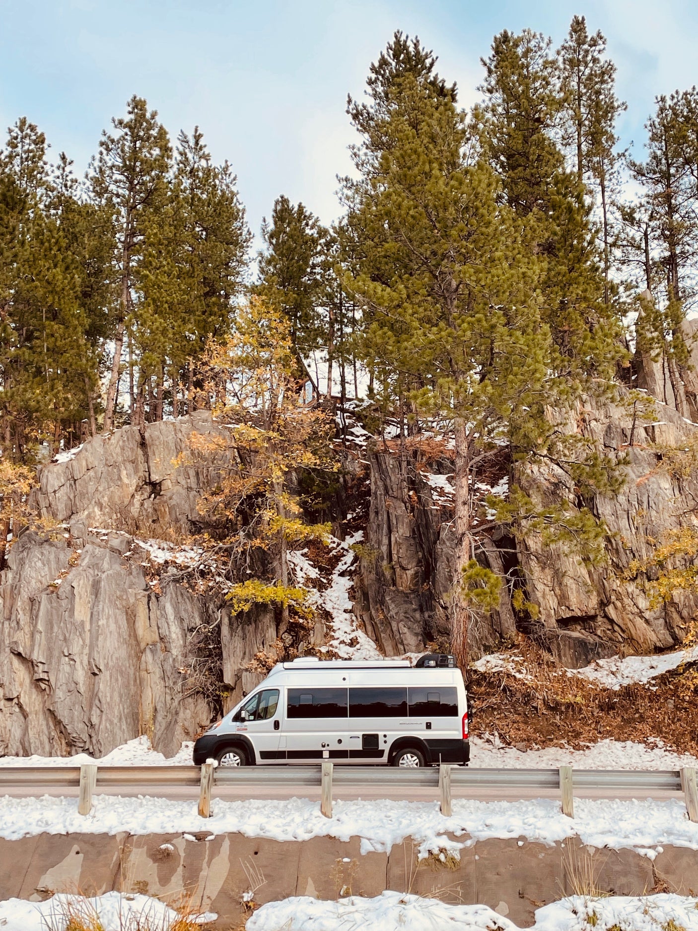 An RV rental, a silver camper van, is parked on a snowy road beside a towering rock face and evergreen trees. Ideal for winter adventures.